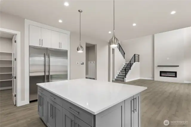 Spacious kitchen with quartz island and built-in refrigerator, offering direct views into the open great room with modern fireplace and sleek staircase. Walk-in pantry and mudroom access add everyday functionality to this elegant design.