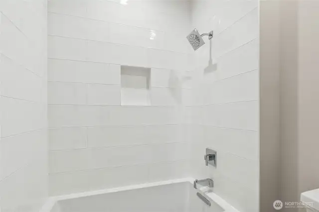 Close-up of the tiled shower and tub surround featuring oversized white subway tile, a built-in shampoo niche, and polished chrome fixtures. Clean, modern design with timeless appeal.