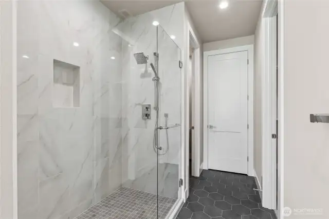 Sophisticated walk-in shower with floor-to-ceiling marble-style tile, frameless glass enclosure, built-in niche, and dual shower heads including a handheld sprayer. Matte hex tile flooring adds a modern touch to this luxurious space.