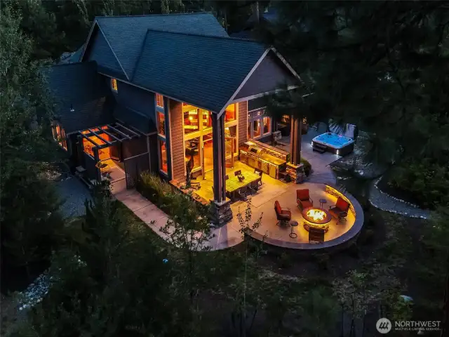 A stunning evening view of the outdoor kitchen, dining table, and firepit gathering area all softly illuminated for cozy evening entertaining. The home’s windows beam with warmth — a true reflection of comfort and lifestyle.