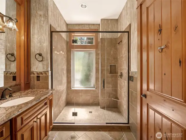 The oversized walk-in shower is beautifully tiled with a frameless glass enclosure, built-in shelving, and natural light streaming through a frosted privacy window.