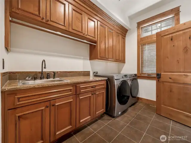 Spacious laundry room includes abundant upper and lower cabinetry, a utility sink with tile backsplash, granite counters, and full-size front-load washer/dryer. A sunny window and thoughtful finishes make laundry day a breeze.