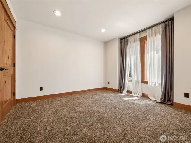Light-filled upper floor guest room or secondary bedroom with warm wood trim, updated carpet, and a wall of windows with soft-filtering curtains — offering peaceful comfort and privacy