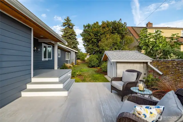 A view of the back yard from the patio, with brick fence running along the property line at right.  One of two storage sheds sits just off the patio.  Covered back deck leads into the mudroom.