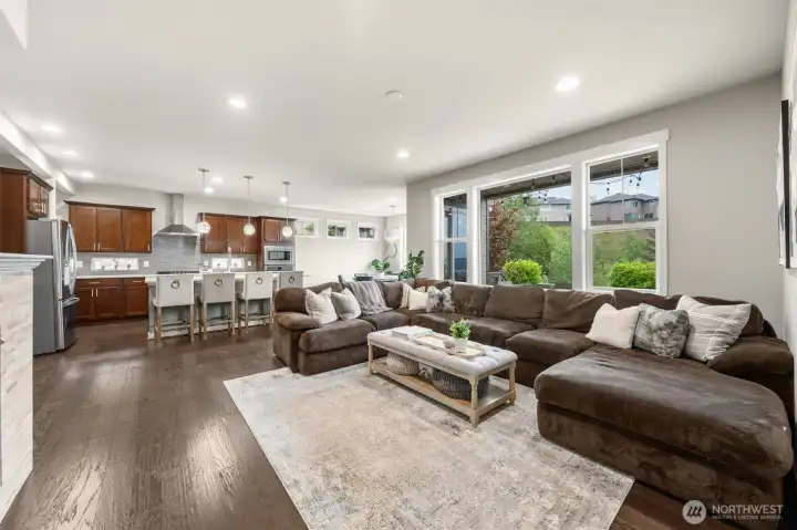 The great room delivers the open floor plan buyers crave — generous seating space flows directly to the kitchen and dining area, all anchored by oversized windows that frame the lush greenery beyond