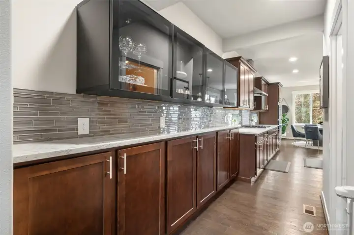 Glass-front display cabinets in a contrasting dark finish add a designer touch to this extended counter run — with a dedicated pantry just to the left offering the storage every home cook dreams of, all flowing seamlessly toward the dining area beyond