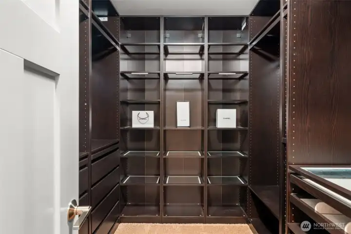 A custom-built walk-in closet with floor-to-ceiling dark wood shelving, glass-front display sections, and built-in drawers — designed for the buyer who takes their storage as seriously as their style.