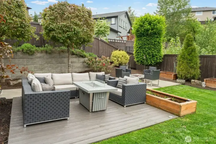 A dedicated outdoor living room complete with a gas fire table, sectional seating, and raised cedar garden beds — all set on a composite deck surrounded by mature landscaping for natural privacy. Every detail of this backyard was designed with intention