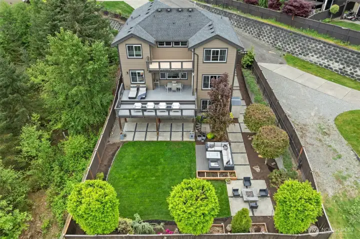 Rarely does outdoor living come this well designed. This aerial view showcases the thoughtfully landscaped backyard featuring multi-level composite decking, designer concrete patio spaces, a fire pit lounge area, raised garden beds, and a lush manicured lawn — all fully fenced for privacy.