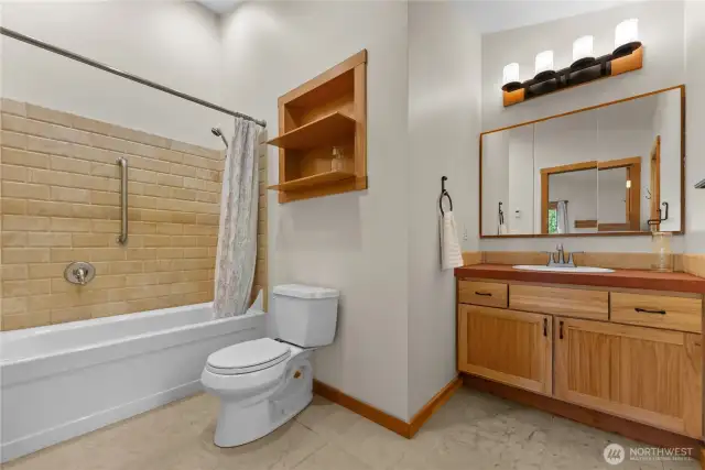 Elegant ensuite bathroom with tile-surround tub/shower combo, built-in shelving, and custom vanity with concrete countertop and warm wood cabinetry.
