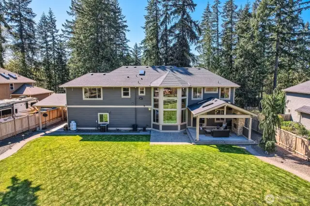 Rear aerial view of the estate home and backyard. A paved walkway with landscape lighting and decorative stone borders encircles the backyard turf, enhancing curb appeal and simplifying mowing and lawn care. Designed for beauty, ease, and year-round enjoyment.