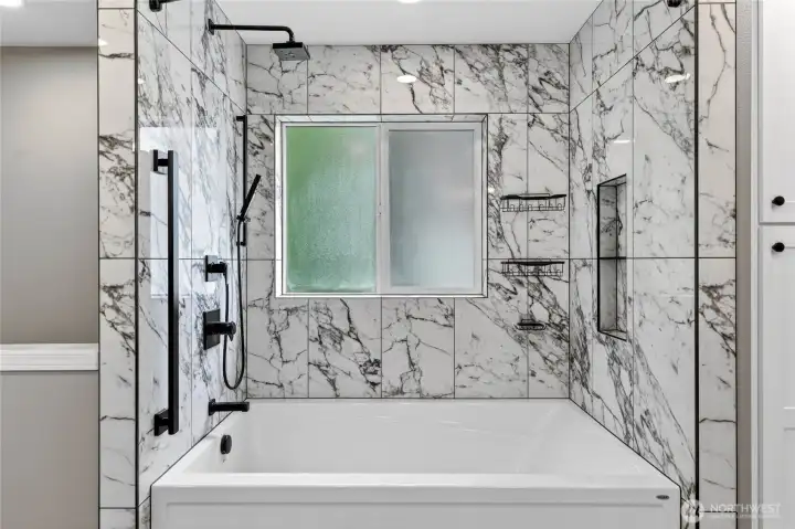 Huge soaking tub with black designer spray wand as well as shower head and grab bar, marble tile surround with toiletry niche