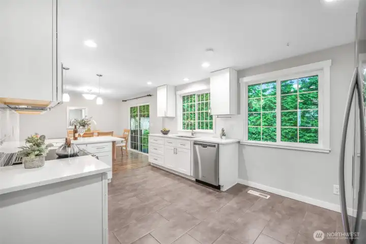 Large windows frame greenery outside, filling the kitchen with natural light. The layout provides both workspace and easy flow to the dining area and outdoor deck.