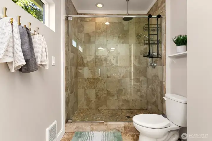 The primary suite features a spacious, walk-in tile shower with frameless glass doors, rainfall and handheld heads, and a pebble stone floor for a true spa-like experience.