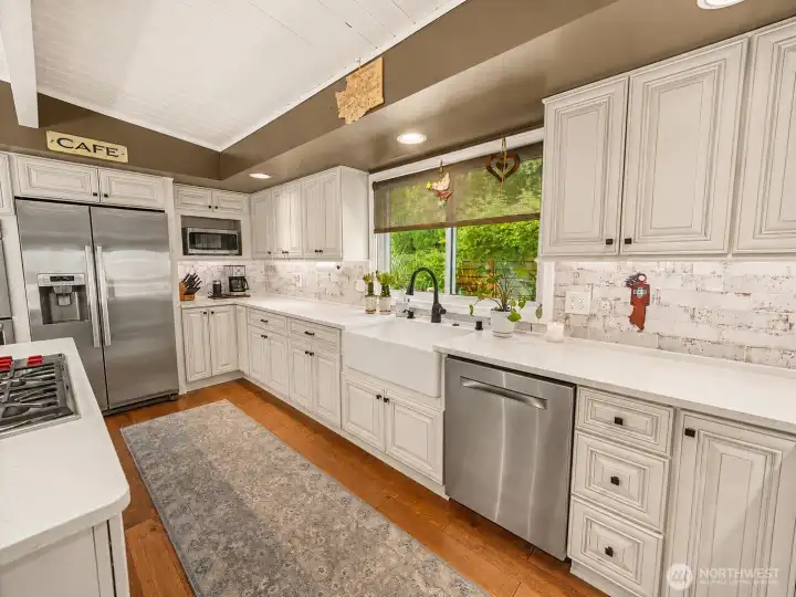 The beautifully updated kitchen blends timeless style with everyday functionality, featuring quartz countertops, custom cabinetry, stainless steel appliances, and a farmhouse sink overlooking lush garden views. Vaulted ceilings and abundant natural light create a bright, welcoming space at the heart of the home.
