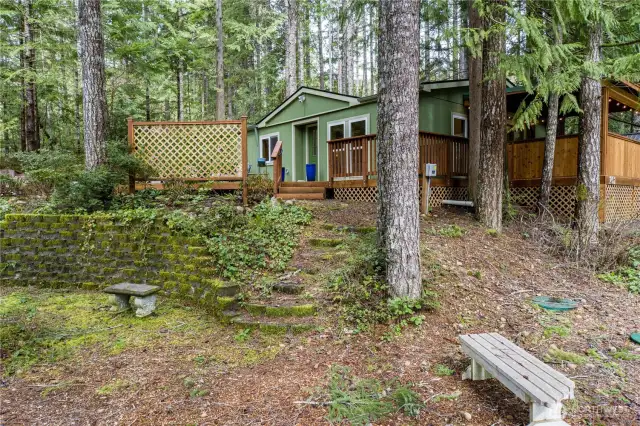 Tucked into the trees, this inviting woodland retreat features a charming front entry with privacy lattice and natural stone steps leading up from a serene garden bench area. Surrounded by lush forest and moss-covered landscaping