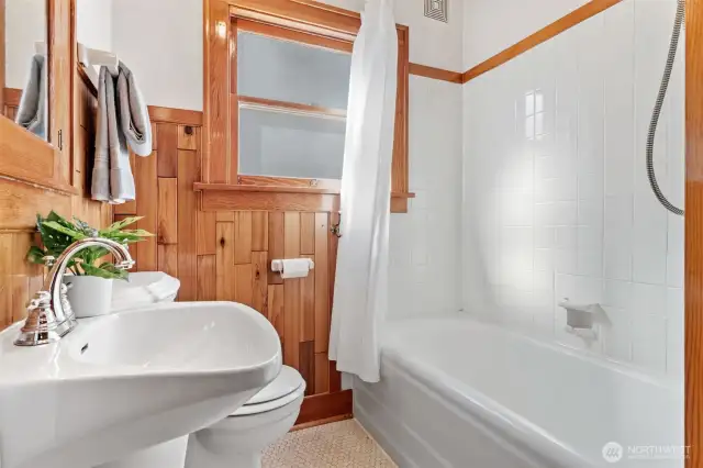 Main-level full bathroom blends timeless charm with modern convenience, featuring classic wood paneling, a pedestal sink, and a tiled shower/tub combo.