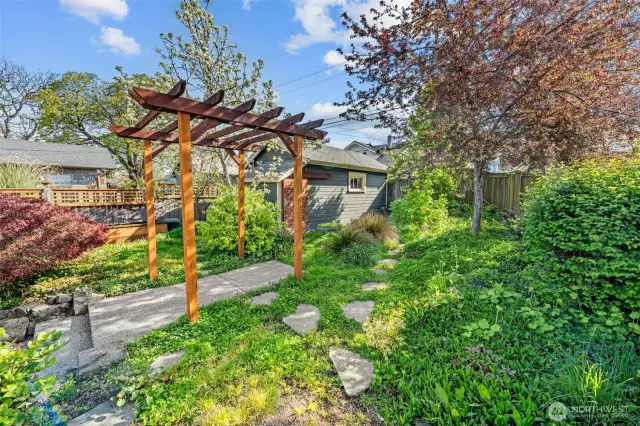 A charming garden path winds beneath a wooden pergola, surrounded by lush greenery and mature trees. Just steps from the back door, this peaceful retreat features apple trees and an Asian pear tree.