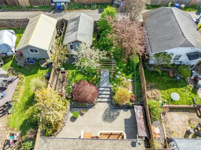 A stunning aerial view of the fully fenced backyard reveals a thoughtfully landscaped garden retreat—complete with tiered stonework, mature trees in full bloom, raised beds, a charming pergola walkway, and plenty of room to relax or entertain in total tranquility.