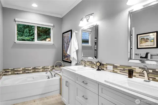 Primary bath. Walk-in closet with top-of the line interior closet organizer system. Heated tile floor. Crown molding. Picture frame beveled mirrors with bell shaped Alabaster style shades for vanity lights above. Double-sink vanity with Designer sinks and Quartz countertop, with Mosaic glass tile backsplash, recessed panel cabinets below. Oval soaking tub with Mosaic glass tile accented backsplash and Quartz counter and window above with two-inch blinds and screen. Paneled Quaker pocket door to watercloset and shower privacy room. Elongated watercloset. Step-in shower with glass doors, handheld shower wand.