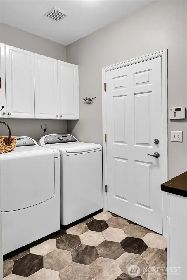 Light, bright laundry room. Paneled Quaker door. Whirlpool® Cabrio Platinum™ dryer and washer stay with home. Paneled white cabinets above. Folding counter with paneled white cabinet above and below. Faux-tile floor. Honeywell® security system. Step down to garage.