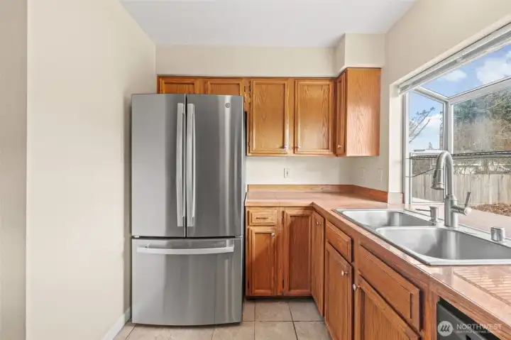 This bright kitchen features rich wood cabinetry paired with a modern stainless steel refrigerator for a perfect mix of style and function. The large garden window above the sink fills the space with natural light and offers a lovely view of the backyard.