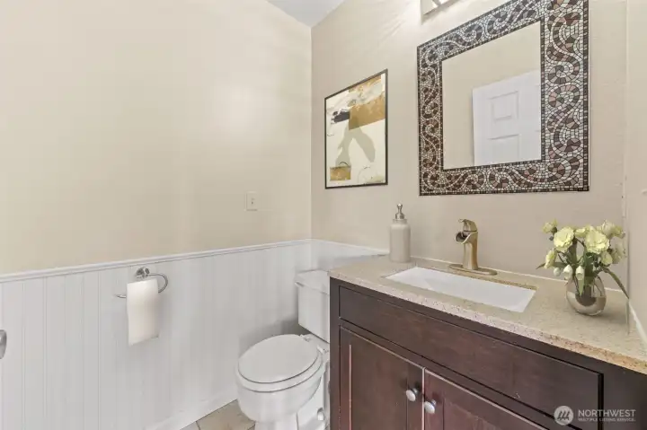 This stylish powder room on the main level features a modern vanity with a stone countertop and elegant mosaic-framed mirror. Crisp wainscoting and soft neutral tones create a clean, polished look that complements the home’s design.