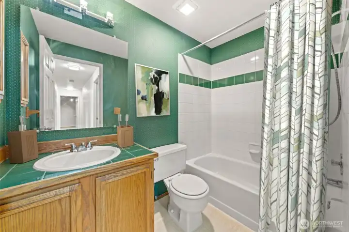 The upstairs full bathroom offers a cheerful and functional layout with classic tile accents and a full shower/tub combination. Warm wood cabinetry and bold green detailing add a touch of character and charm to the space.