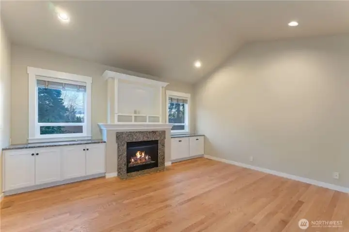 Living room featuring a cozy fireplace with built-in shelving on both sides, vaulted ceilings, and large windows overlooking the private backyard that bring in abundant natural light. Photo depicts the room without furniture; seller furnishings are currently in the home.