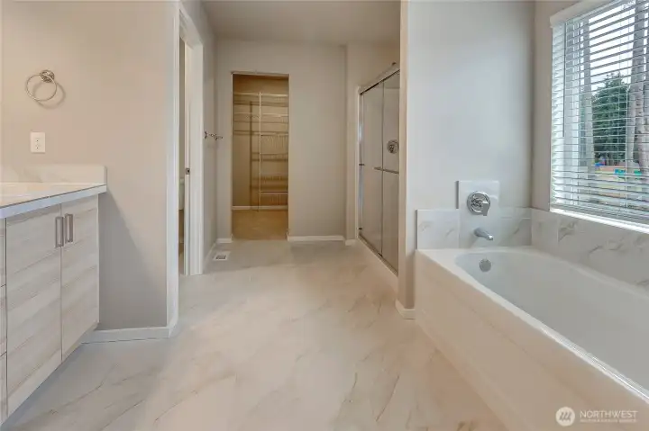 This elegant en-suite bathroom features a beautifully tiled oversized shower with a glass door, a soaking tub, and a double vanity with stylish tile counters. Just beyond, a spacious walk-in closet with wire shelving adds convenience to this bright, luxurious retreat.