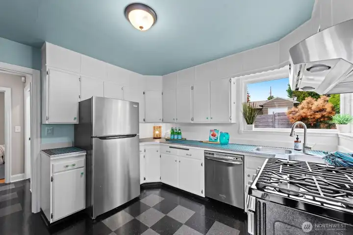 Retro-inspired kitchen with updated stainless steel appliances, checkerboard flooring, and cheerful light blue accents. A large picture window provides ample sunlight and a view of the backyard