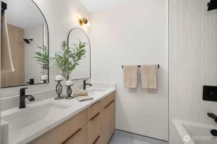 Spa-inspired primary bath with dual sinks, light wood vanity, taj mahal quarts countertops, black fixtures, and elegant designer sconces.