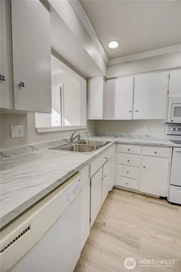 Entrance to kitchen from Living room. Updated countertops with grey veining, shaker-style upper & lower cabinetry with silver knob finishes, and a double basin stainless kitchen sink. Dishwasher located to left of sink near one entrance to kitchen. Sink sits under the pass-through window. Recessed light installed throughout the ceiling for a clean look. NEW LVP floor with a light wood color.