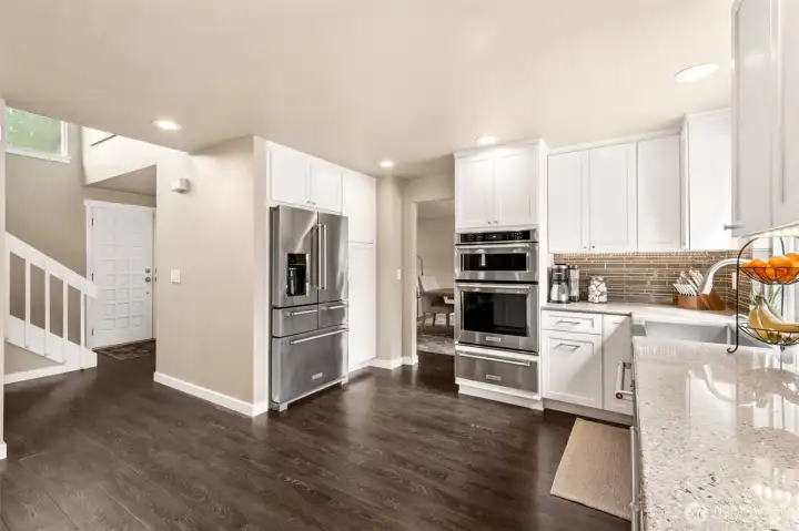 Bright, beautifully remodeled kitchen with crisp white cabinetry, quartz counters, and rich dark flooring creating a warm contemporary contrast. Features include a large farmhouse sink, stainless appliances, gas cooktop island, triple-oven configuration with warming drawer, sleek hardware, and elegant full-height tile backsplash.