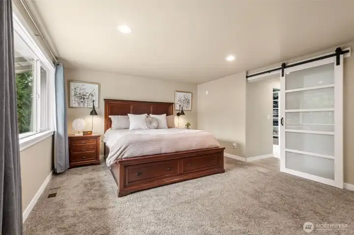 Spacious and serene primary suite featuring plush carpeting, oversized windows for abundant natural light, and a soft neutral palette creating a calm retreat-like atmosphere. The beautifully sized room easily accommodates large furnishings while offering excellent flexibility and comfort. Contemporary frosted glass barn doors add architectural interest and lead to the remodeled ensuite bath and customized walk-in closet, blending modern style with everyday functionality.