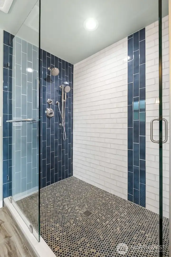Stunning oversized walk-in shower with designer tilework, frameless glass enclosure, pebble-style flooring, and contemporary fixtures including a rainfall showerhead and handheld sprayer. Rich blue accent tile paired with crisp white subway tile creates a fresh, spa-inspired aesthetic with striking modern appeal.