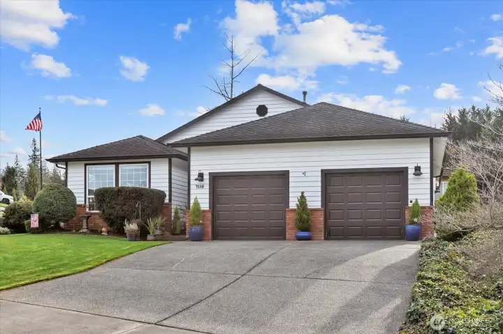 Attractive two-car garage with modern dark doors, framed by lush shrubs and fresh blue planters. Large picture windows overlook the landscaped front yard, showcasing the home’s clean lines and well-kept exterior. A spacious driveway offers ample parking.