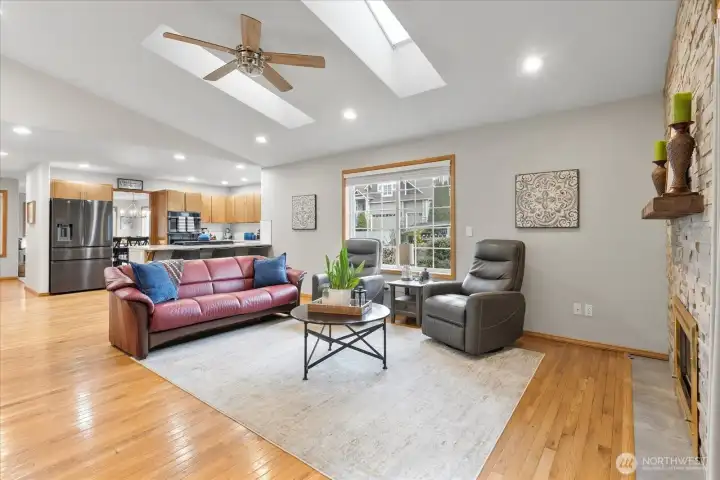 Open-concept living area with seamless flow into the kitchen and dining spaces. Highlights include hardwood floors, skylights, a ceiling fan, and a cozy fireplace.