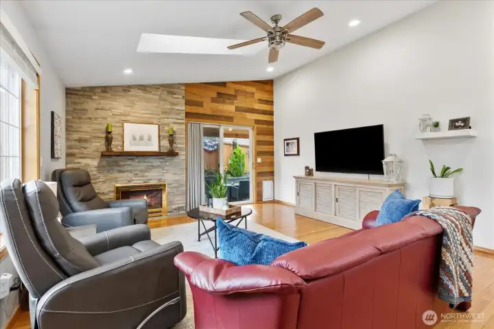 Inviting living area featuring a stone-surround fireplace, hardwood flooring, and overhead skylights that bring in natural light. Sliding glass door provides access to the outdoor space.