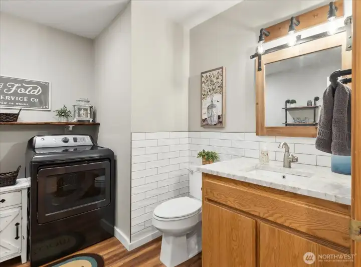 Combined bathroom and laundry area with vanity, quartzite countertop, and front-loading dryer. Open shelving provides additional storage.
