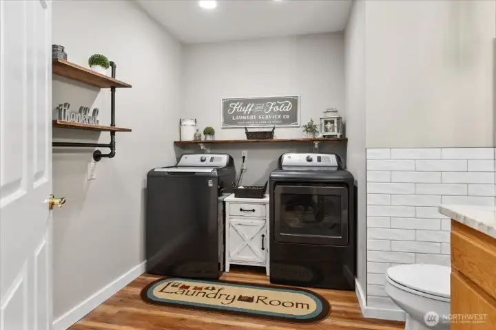 Laundry room with washer, dryer, and wood shelving. Decorative accents and overhead lighting complete the space.
