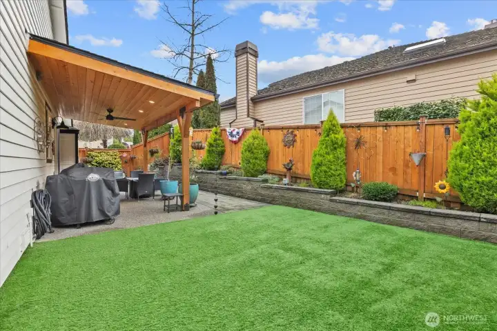 Patio area with seating space, potted plants, and raised garden beds along the fence line. Artificial turf and stone accents create a low-maintenance yard.