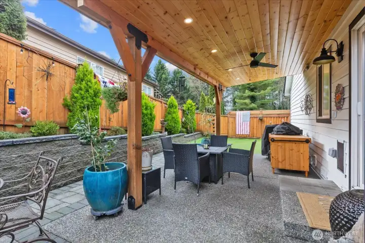 Covered outdoor dining area with ceiling fan and lighting. Surrounding garden beds and fence décor add visual interest.