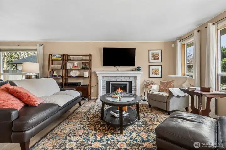 There’s something very satisfying about a living room that feels balanced the second you walk in. The brick wrapped gas fireplace anchors the space with a bit of texture and personality, topped by a classic white mantle that feels timeless instead of trendy. The TV mount above makes the setup effortless, no measuring tape drama required. Sunlight filters through the pair of front windows and that larger picture window near the dining area, giving the room a soft, lived in glow throughout the day. Wrapped in those creamy latte toned walls, the whole space feels warm, inviting, and ready to host everything from quiet evenings to full house gatherings.