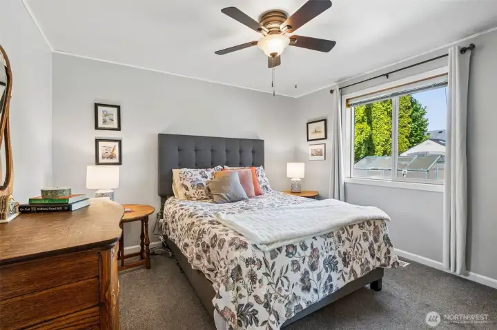 Bedroom two keeps the mood calm and comfortable with that soft mushroom toned carpet underfoot and the prettiest whisper of dove gray on the walls. A ceiling fan helps keep the air moving year round, whether it is warm summer evenings or cozy winter nights. The window looks out over the backyard where you can catch a peek of the greenhouse roof and all that green space beyond. And because this is true one story living, every bedroom sits on the same level, which makes daily routines feel simple and accessible for guests, family, or anyone thinking long term.