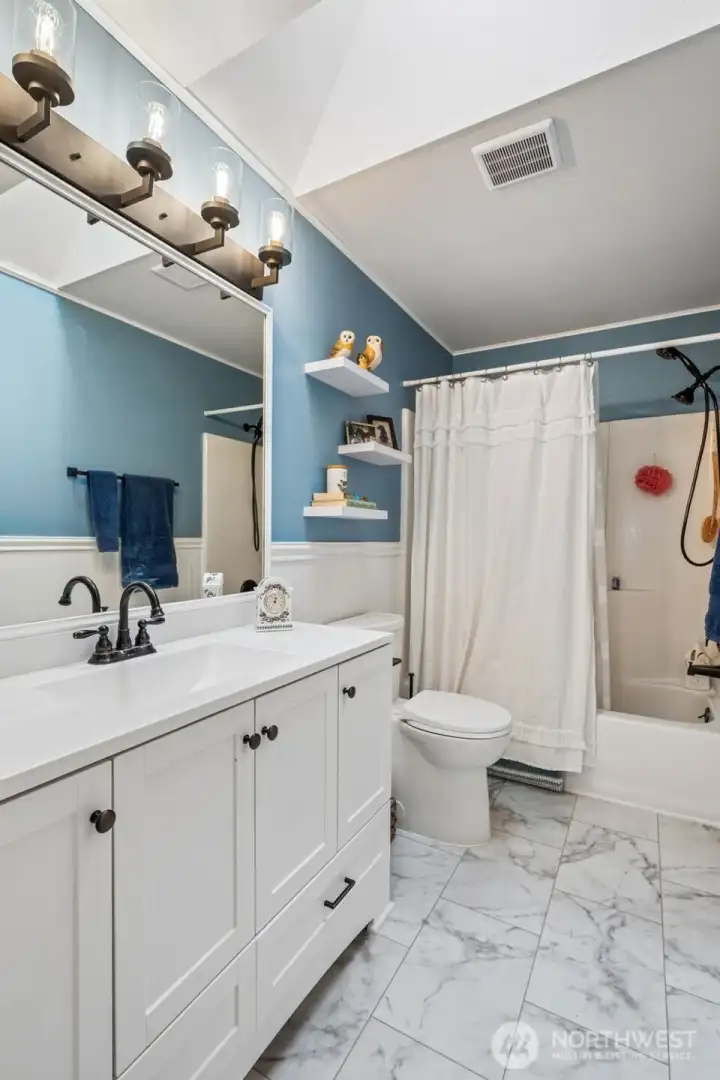 The main bathroom leans classic with a little personality twist. Beadboard wainscoting grounds the space while the soft stormy blue walls bring just enough mood to keep it interesting. A long vanity gives everyone elbow room for those busy morning routines, and the full tub setup features a dual showerhead with handheld sprayer for extra flexibility. The sleek skirted style toilet makes cleaning feel less like a chore, and overall the room lands in that sweet spot of practical and polished.