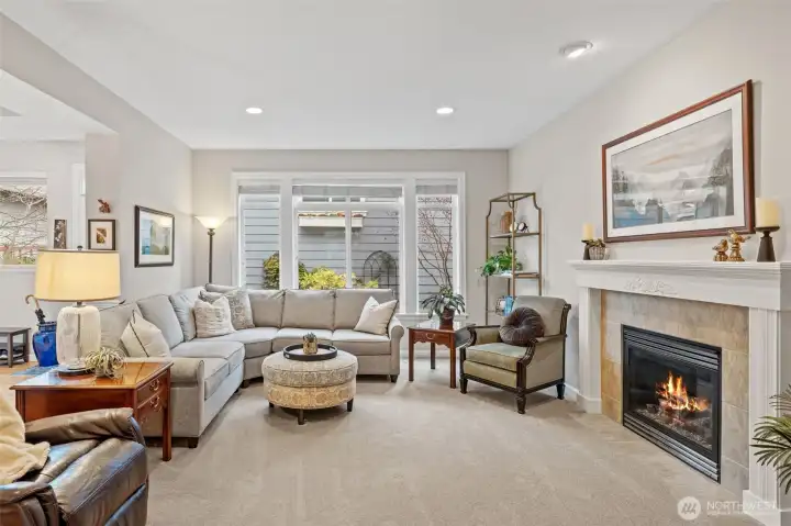 The living room feels bright and inviting with large windows that draw in natural light and frame the gardens outside. The gas fireplace is elevated by a beautifully detailed mantel and a recessed art light above, creating a refined focal point that anchors the entire space.