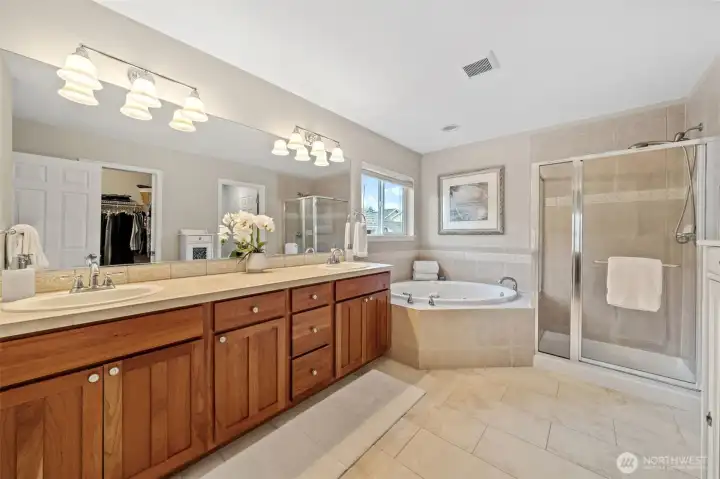 The Primary Suite bath offers a spacious, spa-like layout with a large dual-sink vanity and expansive mirror that brightens the entire room. A soaking jetted tub and separate glass-enclosed shower create a relaxing retreat, complemented by warm tile finishes and ample storage throughout.
