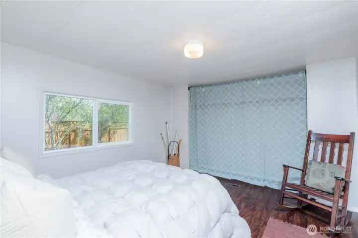 Comfortable and spacious primary bedroom featuring warm wood-tone flooring, a large window for natural light, and a neutral color palette ready for your personal style. The generous layout offers flexibility for larger furniture arrangements, while the closet area provides practical storage. A peaceful retreat with views of the private backyard and mature landscaping beyond.