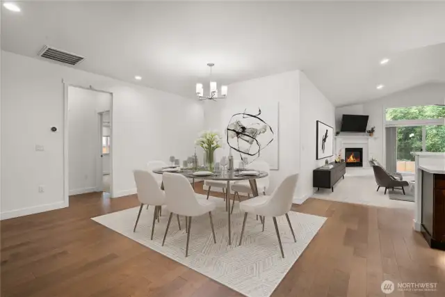 Spacious open-concept living and dining area featuring warm hardwood floors, modern lighting, and a cozy fireplace with backyard views. Virtually staged.